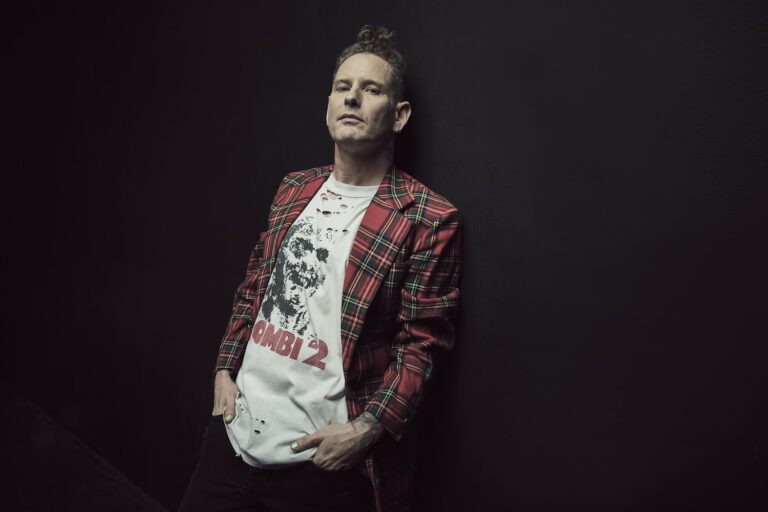 Corey Taylor leans against a black backdrop, shown from just below the waist up, wearing dark pants, white graphic tee, and an open red plaid jacket.