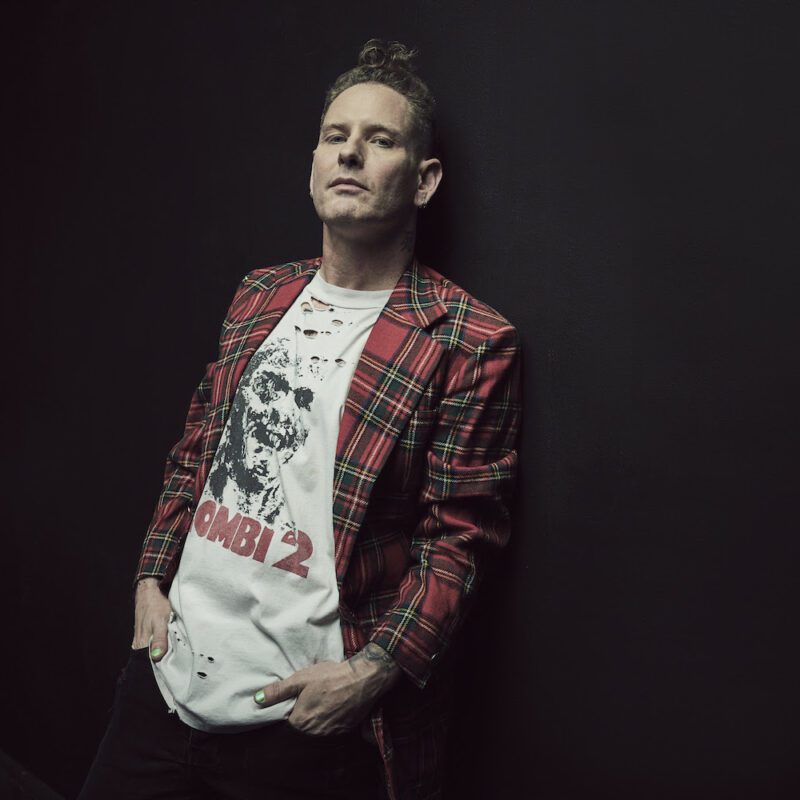 Corey Taylor leans against a black backdrop, shown from just below the waist up, wearing dark pants, white graphic tee, and an open red plaid jacket.